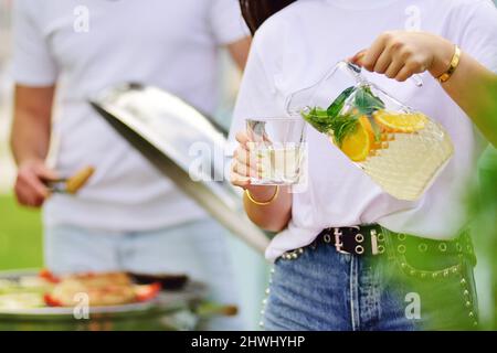 une jeune femme verse de la limonade fraîche d'un décanter sur le fond d'un homme qui rôde un barbecue dans la nature Banque D'Images