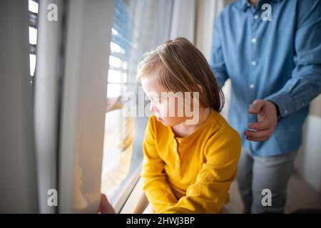Triste petite fille avec le syndrome de Down regardant par la fenêtre avec son père à l'arrière-plan. Banque D'Images