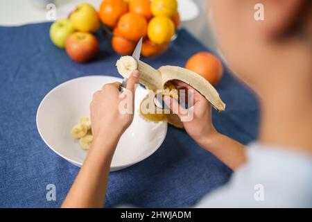 Gros plan sur les mains d'une femme caucasienne inconnue coupant la banane par la table à la maison - vue arrière haut angle Copy space - concept de saine alimentation Banque D'Images