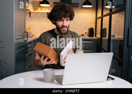 Portrait de heureux surpris jeune homme mauriquement ouverture boîte cadeau avec le présent pendant l'appel vidéo sur ordinateur portable assis à une table dans la cuisine. Banque D'Images