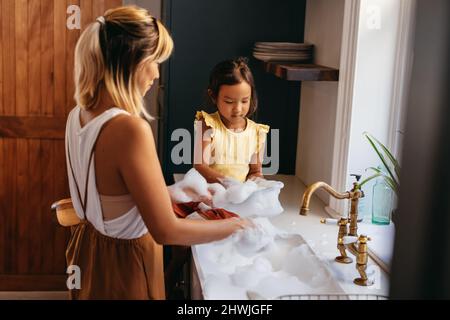 Petite fille apprenant à laver la vaisselle à la maison. Adorable petite fille aidant sa mère avec les plats tout en étant assis sur le comptoir du lavabo. Mère Banque D'Images