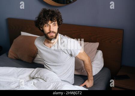 Portrait d'un jeune homme beau assis sur le lit après s'être réveillé et avoir regardé le matin dans la fenêtre. Banque D'Images