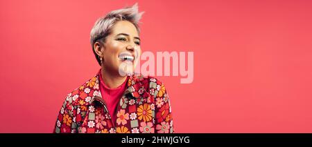 Gaie femme d'hipster regardant loin avec un sourire sur son visage dans un studio. Une jeune femme heureuse debout seule sur fond rouge. Jeune à la mode Banque D'Images