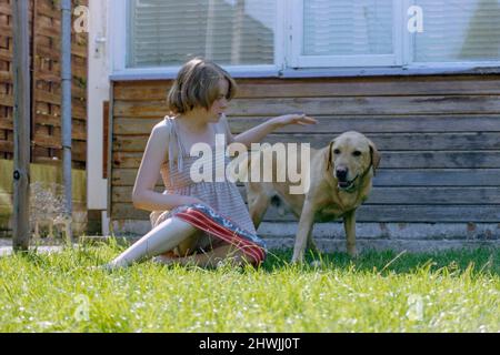 jolie jeune femme avec bob blond coupe de cheveux et robe d'été légère à la mode au début de 1980s pats son chien dans le jardin rugby angleterre royaume-uni Banque D'Images