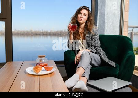 Femme dans une atmosphère chaleureuse, petit déjeuner avant travail à distance, assis dans un fauteuil près de la fenêtre panoramique et table avec thé de fruits et croissant, tenir la tasse Banque D'Images