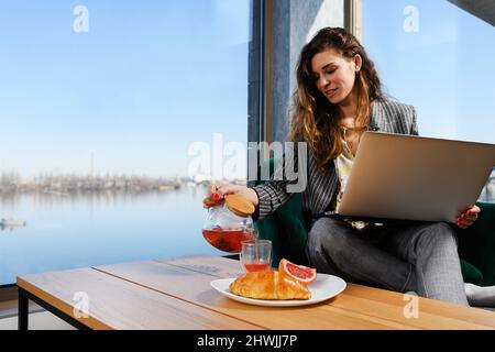 Jeune femme d'affaires, indépendant assis avec un ordinateur portable avec un intérieur agréable, vue sur la rivière dans la fenêtre panoramique et le petit déjeuner thé, croissant et pamplemousse Banque D'Images