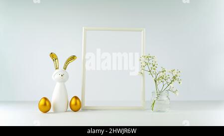 Décor de Pâques sur le fond d'une table et d'un mur blancs. Œufs et un lapin aux oreilles dorées. Cadre et fleurs. Maquette minimaliste pour votre texte. Bann Banque D'Images