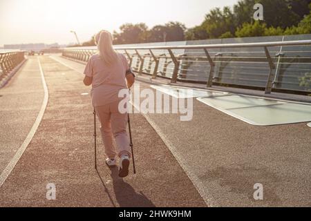 Sportive mature lady pratique la marche nordique sur une passerelle vide au coucher du soleil Banque D'Images