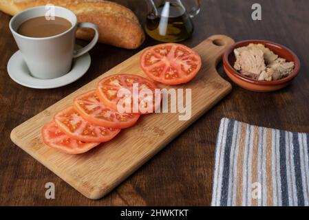 Des tranches de tomate sur une planche en bois à côté d'un café avec du lait, du pain et une assiette avec du thon pour préparer un toast. Banque D'Images