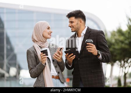 Un couple d'affaires arabe s'est amusé et a parlé ensemble en marchant dans la rue pendant une pause-café. Femme en hijab montrant son écran de smartphone à son collègue, partageant une idée ou des nouvelles Banque D'Images