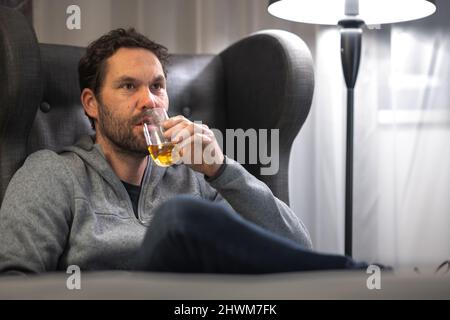 Le jeune hipster est sans expression et il boit une boisson alcoolisée dans un verre dans une chambre d'hôtel. Banque D'Images