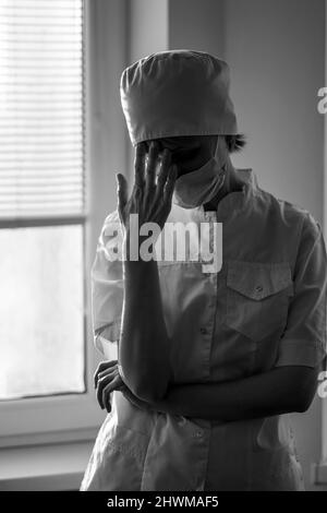 Une femme fatiguée médecin se tient près de la fenêtre. Photo en noir et blanc. Banque D'Images