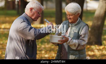 Genre caucasien retraité gris-cheveux homme aide les gens dans le besoin en partageant sa soupe maison avec la femme âgée sans abri. Photo de haute qualité Banque D'Images