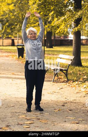 Photo verticale d'une femme âgée à la retraite aux cheveux gris qui étire ses bras dans un parc public, en profitant de l'air frais et du beau temps d'automne. Photo de haute qualité Banque D'Images