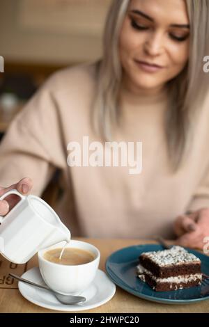 Photo verticale de la femme blonde souriante floue versant de la crème de lait dans le café, mangeant un gâteau au brownie. Café vacances plaisir Banque D'Images