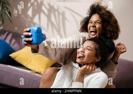 Deux jeunes amies africaines heureux prenant des selfies avec un appareil photo instantané assis sur un canapé à la maison Banque D'Images