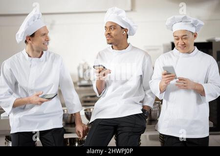 Trois cuisiniers multiraciaux parlent en se tenant debout avec un smartphone pendant une pause dans la cuisine du restaurant. Hommes asiatiques, latins et européens en uniforme blanc pendant une pause au travail Banque D'Images