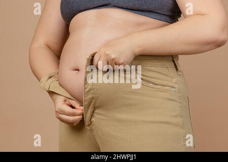 Gros plan de la femme ne peut pas mettre des jeans en raison de la flabby grand abdomen sur fond beige. Graisse viscérale. Corps positif. Gain de poids soudain. Serré Banque D'Images