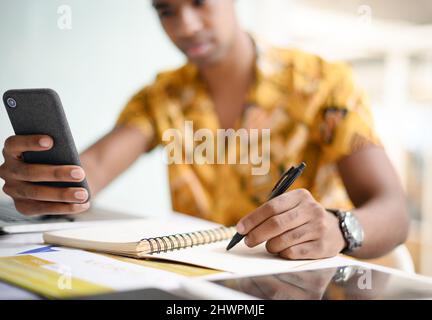 Je dois enregistrer ces informations. Gros plan d'un homme d'affaires méconnu qui écrit des notes en utilisant un téléphone portable dans un bureau. Banque D'Images