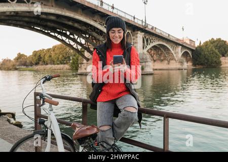 Jeune femme souriante à vélo utilisant un smartphone assis sur un garde-corps Banque D'Images