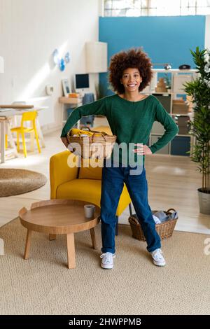 Femme afro souriante avec main sur la hanche tenant le panier dans le salon Banque D'Images