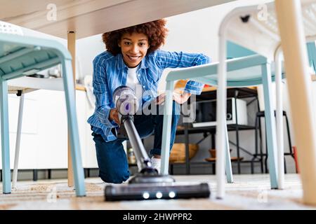 Une jeune femme souriante se caroupe à l'aide d'un aspirateur sous une table à manger à la maison Banque D'Images