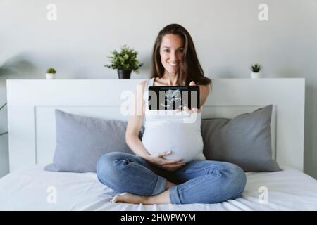 Femme enceinte souriante montrant une échographie au moyen d'un smartphone à la maison Banque D'Images