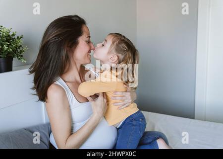 Jolie fille embrassant le nez de la mère enceinte sur le lit à la maison Banque D'Images