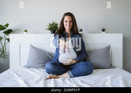 Femme enceinte souriante montrant des chaussons de bébé sur le lit Banque D'Images