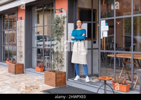 Propriétaire de café souriant debout avec les armes croisées dans la porte Banque D'Images