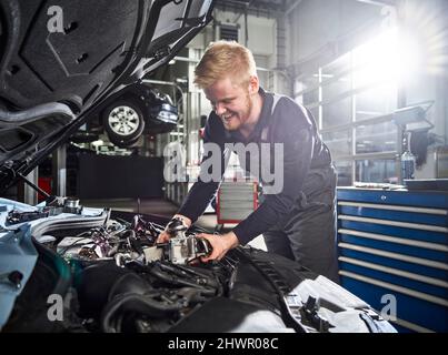 Jeune homme blond souriant mécanicien répare le moteur de voiture dans l'atelier automobile Banque D'Images