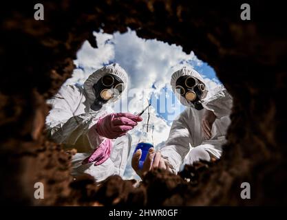 Deux travailleurs de laboratoire travaillant dans des vêtements de protection, des gants en caoutchouc et des respirateurs préchargés de prélever un échantillon de la teneur en sol de la terre dans un flacon contenant un réactif à des fins de recherche. Vue de l'intérieur du puits. Banque D'Images