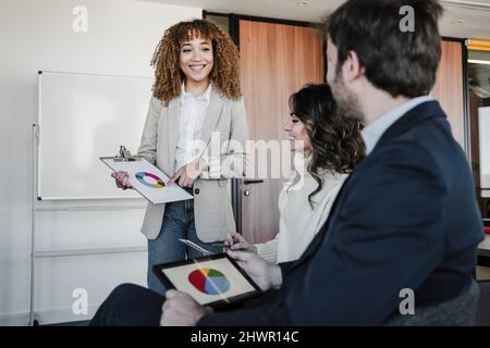 Une jeune femme d'affaires souriante explique le graphique à secteurs sur le presse-papiers aux collègues du bureau Banque D'Images