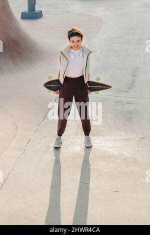 Femme souriante tenant une planche à roulettes sur une rampe de sport dans le parc Banque D'Images