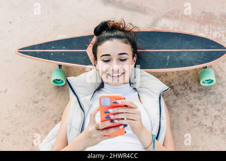 Femme souriante couchée sur un skateboard à l'aide d'un smartphone dans un parc Banque D'Images