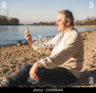 Joyeux homme senior qui passe des appels vidéo par téléphone portable à la plage par beau temps Banque D'Images