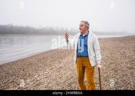 Homme senior effectuant des appels vidéo via un smartphone à la plage Banque D'Images