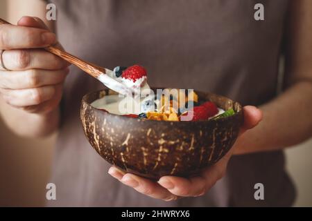 Femme mangeant un petit déjeuner sain dans un bol de noix de coco. Vaisselle écologique et sans gaspillage. Yaourt aux flocons de maïs et aux fruits rouges Banque D'Images