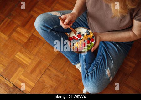 Petit déjeuner sain à la maison. Femme assise sur le sol et mangeant du yaourt avec des fruits et des flocons de maïs. Plats végétariens Banque D'Images