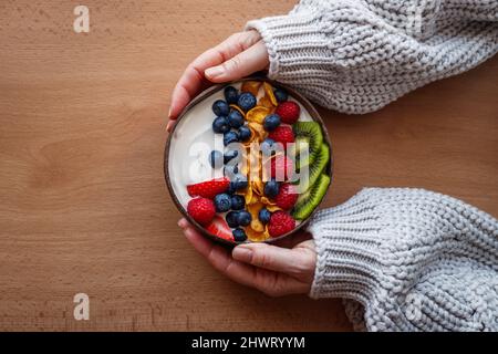 Petit déjeuner sain dans un bol de noix de coco sur une table en bois. Yaourt aux flocons de maïs et aux bleuets, aux fraises, aux kiwis et aux framboises. Chandail à la main pour femmes. Banque D'Images