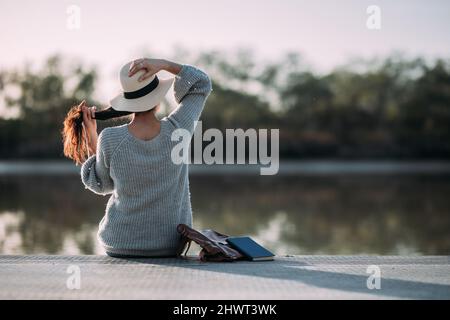 Jeune femme méconnaissable mettant un chapeau en pleine nature, assise sur un quai au bord d'une rivière Banque D'Images