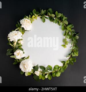 Couronne à cadre rond avec fleur de roses blanches et branches sur fond noir. Concept floral minimaliste. Pose à plat. Copier l'espace. Banque D'Images