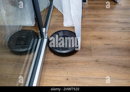 Un aspirateur robot moderne nettoie les panneaux en vinyle près de la fenêtre du patio, un robot de nettoyage autonome. Banque D'Images