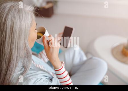 Femme buvant du thé et utilisant le téléphone portable à la maison Banque D'Images