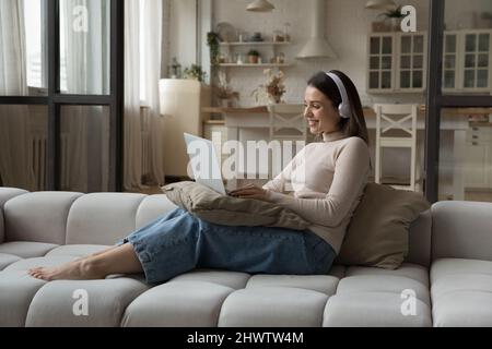Une jeune femme souriante dans un casque utilise un ordinateur portable assis sur un canapé Banque D'Images