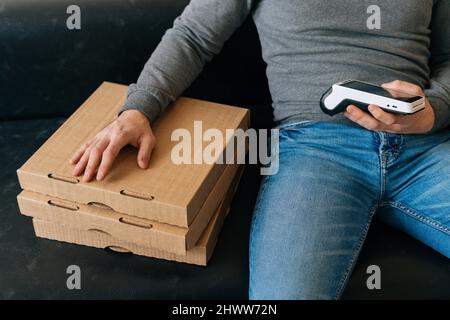 Gros plan d'un livreur méconnaissable qui tient le terminal sans fil de point de vente de paiement en attente du client, assis sur un canapé avec des boîtes de pizza dans le hall Banque D'Images