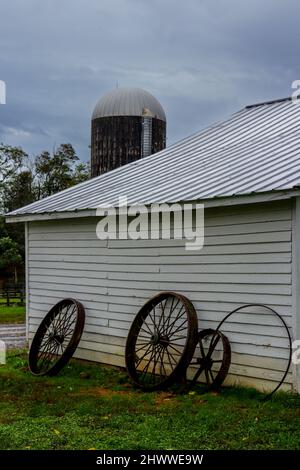 Vieux, rouillé roues de chariot, se pencher contre un bâtiment blanc de ferme avec un silo en arrière-plan. Banque D'Images