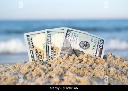 Trois billets de cent dollars à moitié ensevelis dans le sable sur un bord de mer sablonneux Banque D'Images