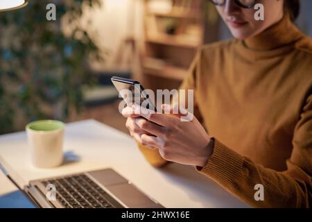 Gros plan sur une jeune femme d'affaires qui tape un message sur un téléphone mobile tout en étant assise sur son lieu de travail avec un ordinateur portable au bureau Banque D'Images
