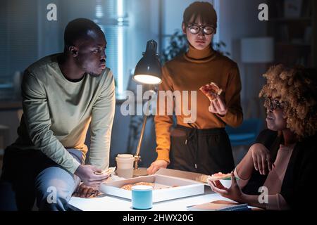 Groupe de gens d'affaires mangeant de la pizza ensemble à la table pendant leur travail d'équipe au bureau jusqu'à tard dans la nuit Banque D'Images